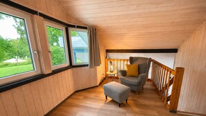 A room with a wooden ceiling, a grey sofa with a yellow cushion, a small ottoman, and a window with curtains. - Skandinavisches Ferienhaus mit Privatsauna im Hafendorf Muritz (Mirow)
