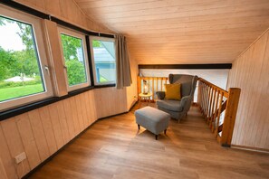 Ein Zimmer mit Holzbalkendecke, einem grauen Sofa mit gelbem Kissen, einem kleinen Hocker und einem Fenster mit Vorhängen.