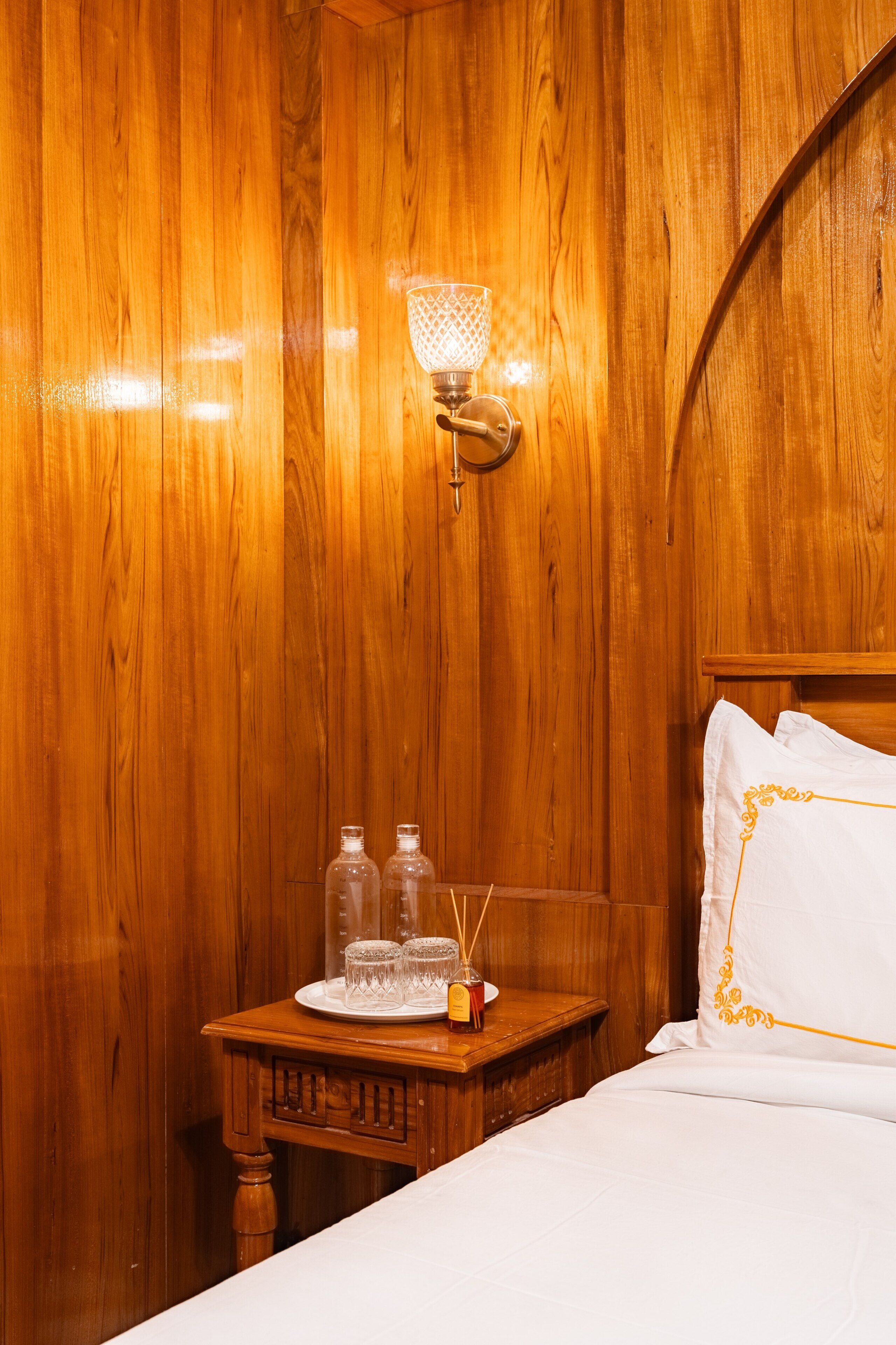 Een kamer met houten lambrisering, een bed, een nachtkastje met lamp en een dienblad met glazen en een fles.