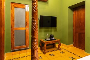 A room with a wooden door, a television mounted on the wall, and a small table with a coffee maker. - Hotel Palais De Versailles (Puducherry)