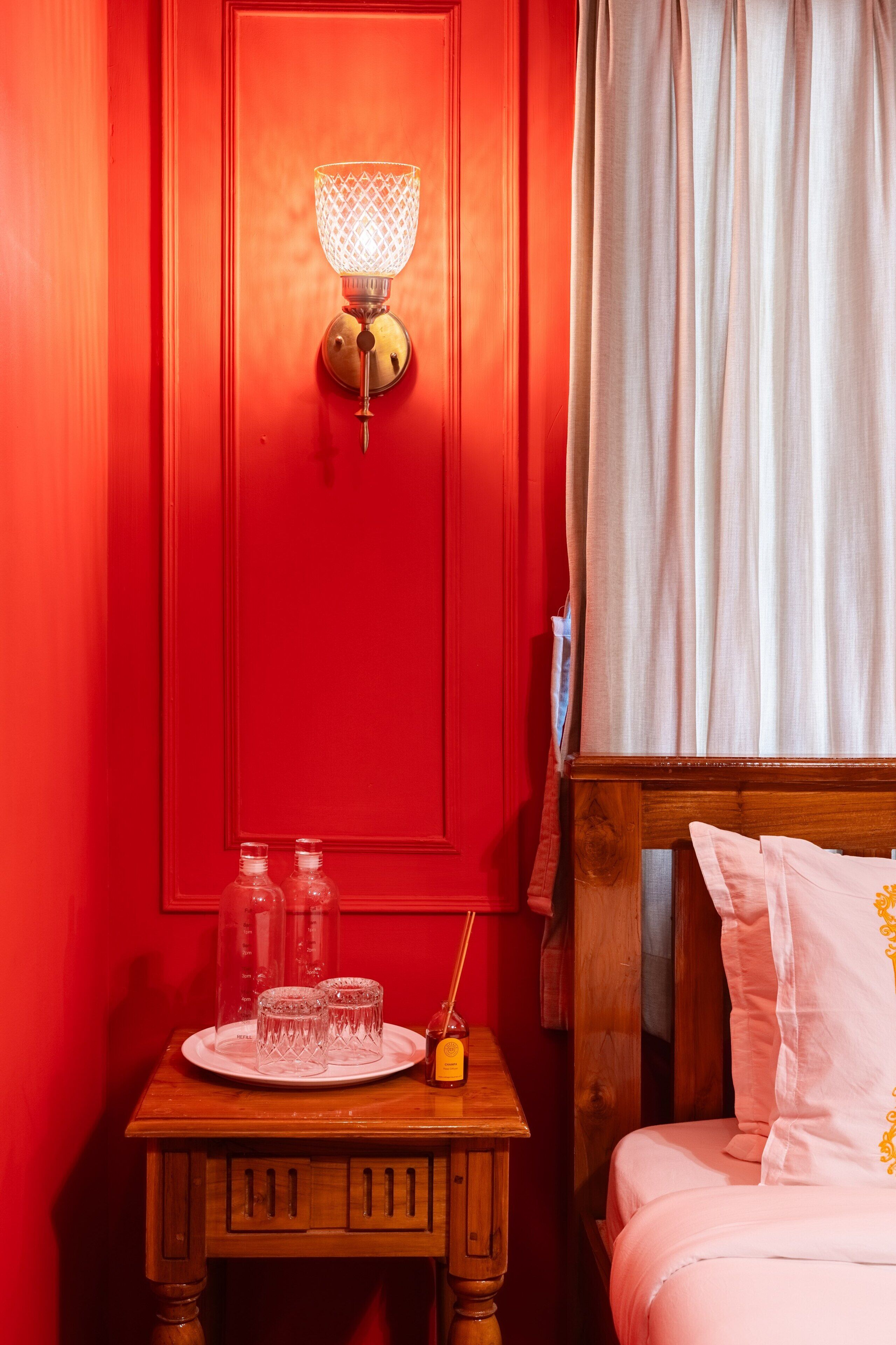 A red door with a wall-mounted light fixture, a bedside table with a glass candle holder, and a bed with white bedding.