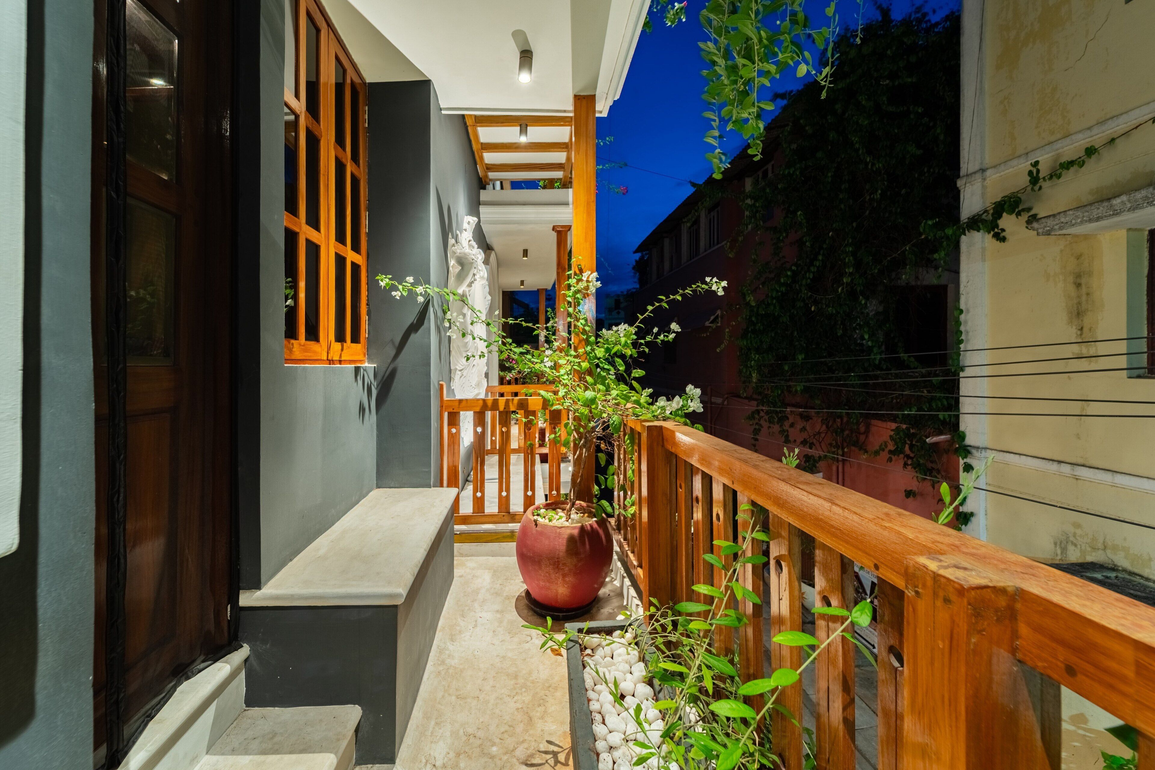A narrow outdoor corridor with a wooden railing, a potted plant, and a staircase leading to an upper level.
