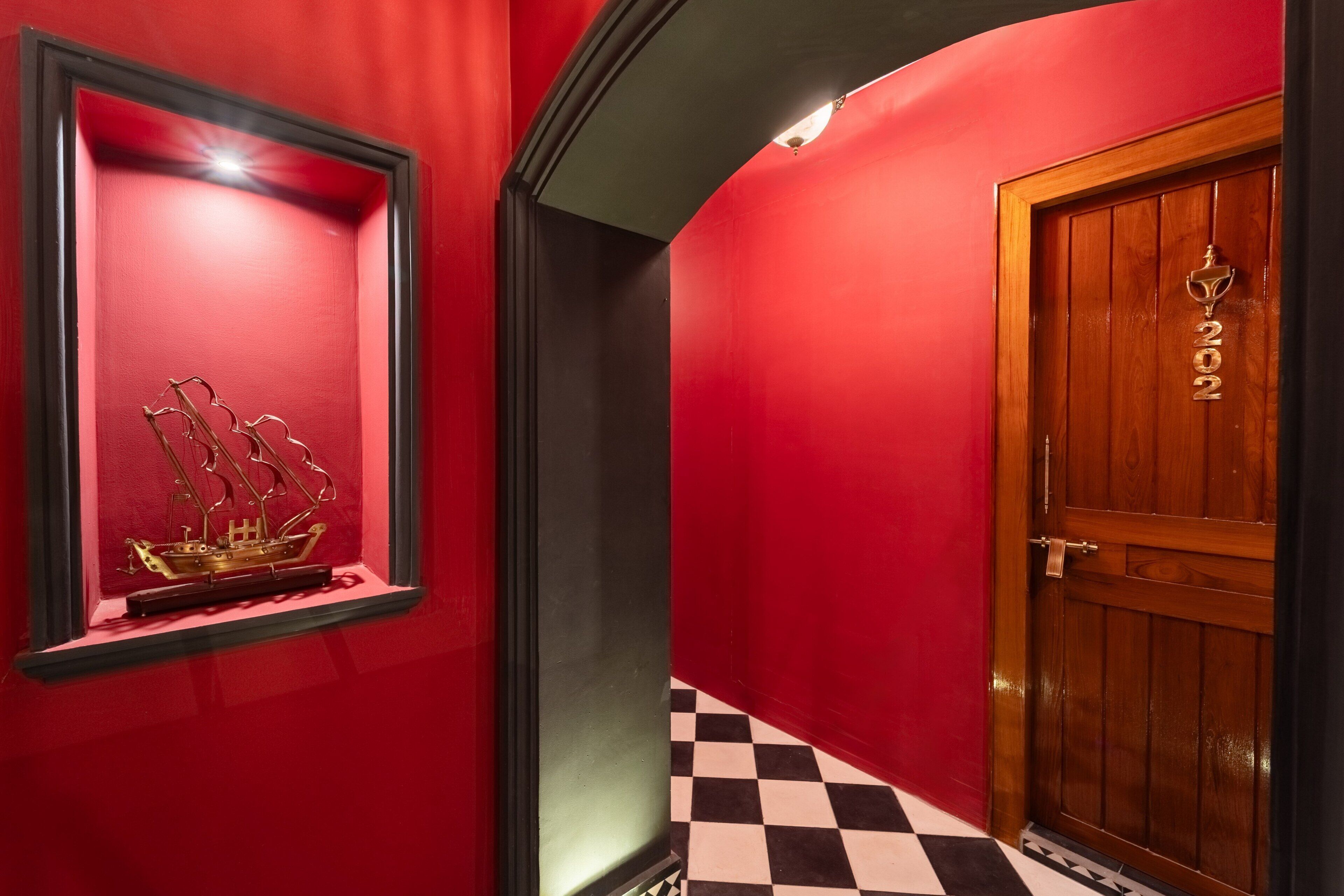 A hallway with a red wall, a framed ship model, a checkered floor, and a wooden door with a number.