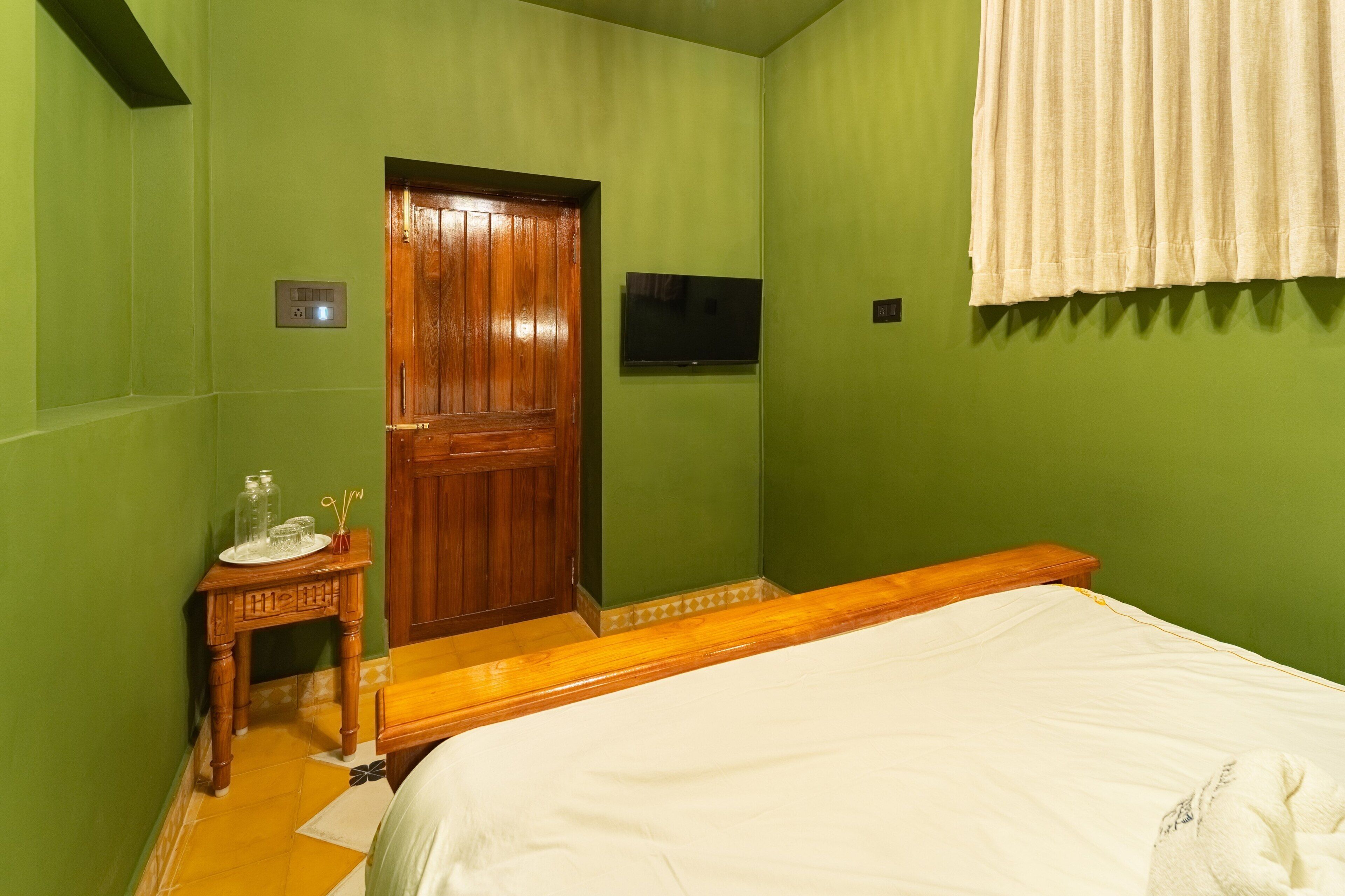 A room with a bed, a wooden door, a small table with a glass jar and spoon, and a television on the wall.