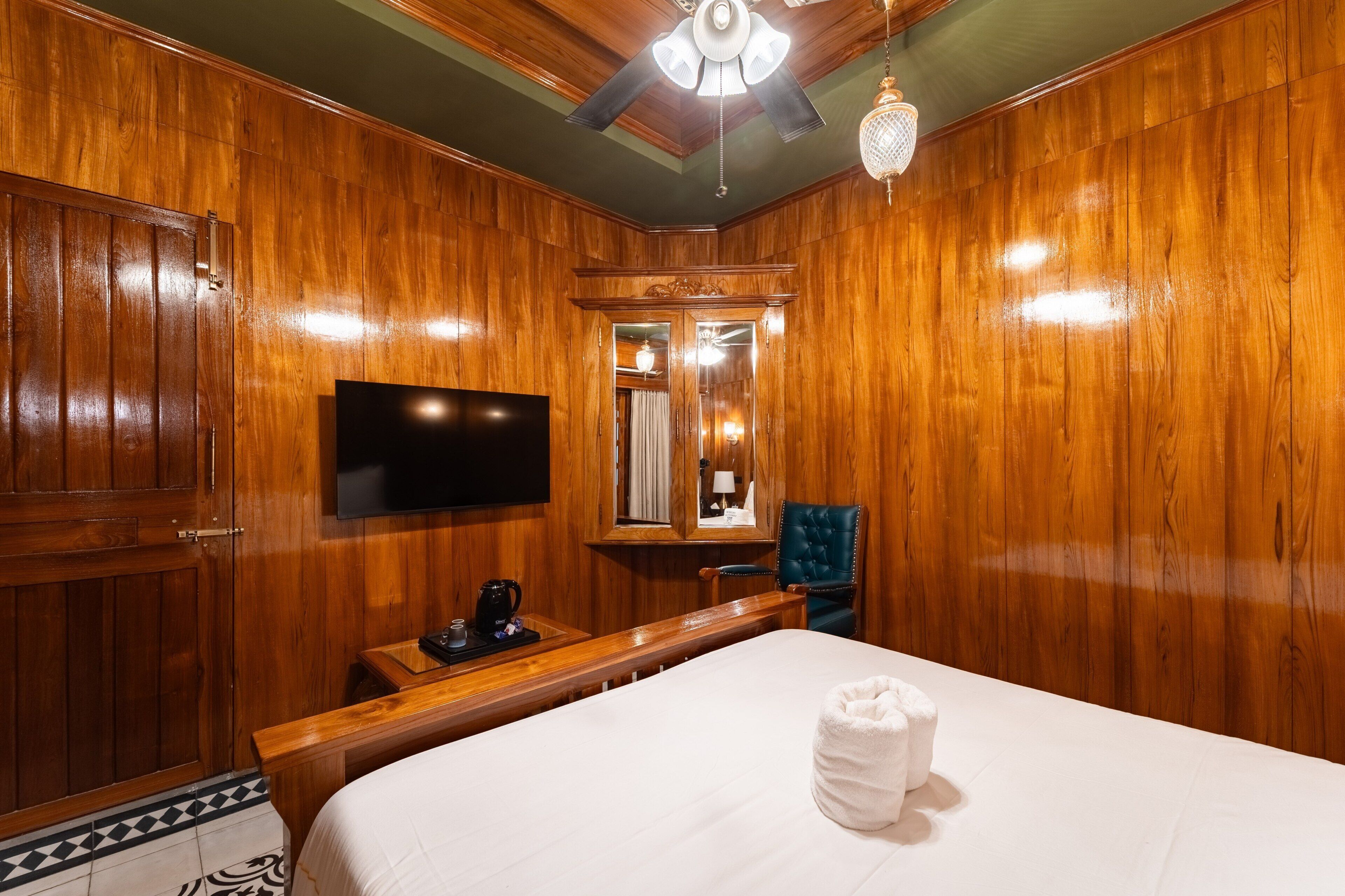 Habitación de hotel con paneles de madera, televisor de pantalla plana, cama con edredón blanco, cafetera, una silla y un espejo.