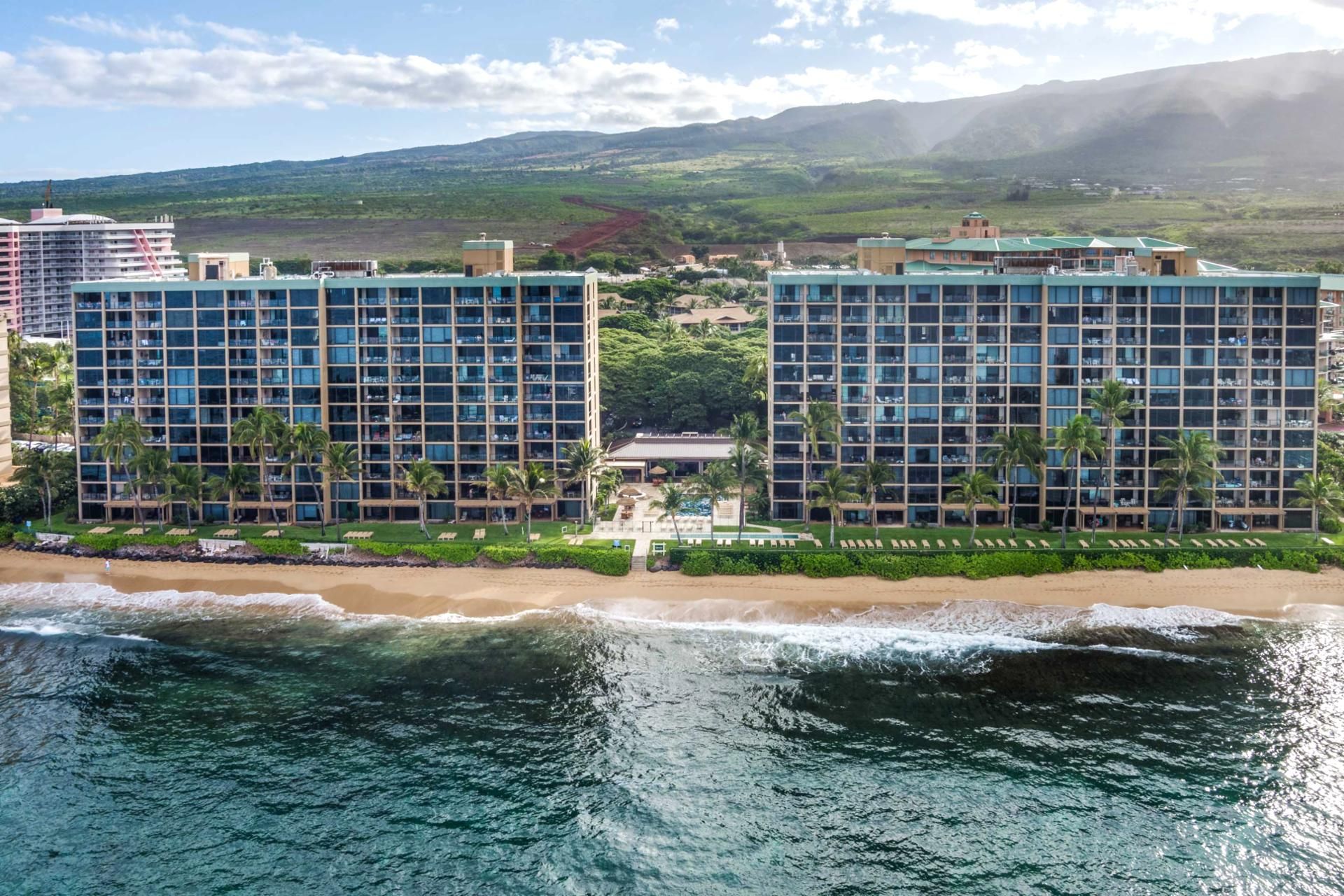 Un hotel frente a la playa con varios edificios de gran altura, una zona de piscina y vistas al océano y a las montañas.