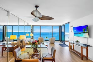 Un salon moderne avec une grande table en verre, des sièges confortables, un ventilateur de plafond et un téléviseur diffusant une scène de plage tropicale.