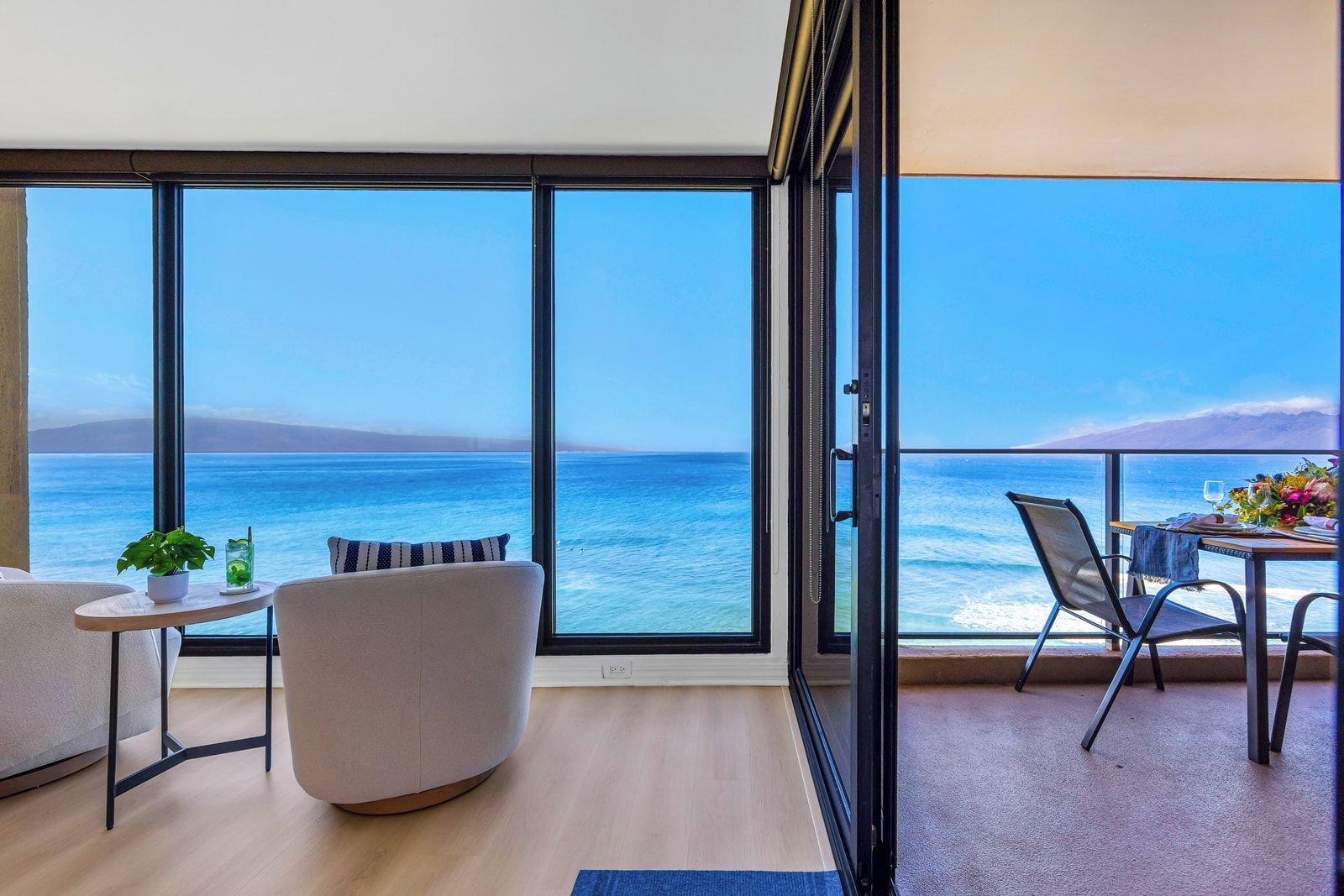 A balcony with a view of the sea, a dining table with chairs, a sofa, and a small table with a plant and drink.