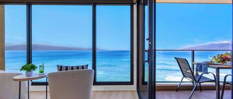 A balcony with a view of the sea, a dining table with chairs, a sofa, and a small table with a plant and drink.