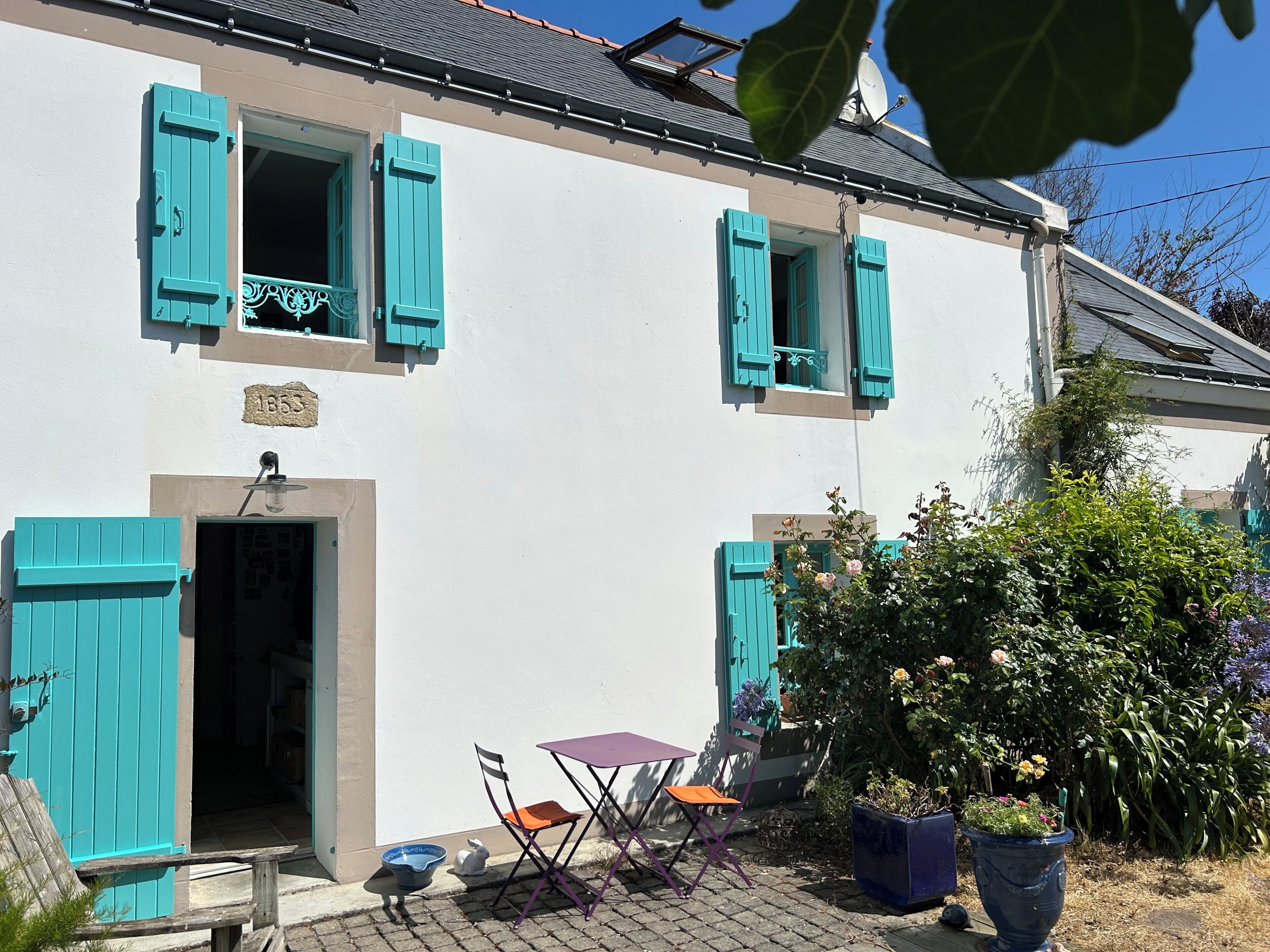 Un bâtiment blanc avec des volets turquoise, un petit espace extérieur aménagé avec une table et des chaises, et un pot de fleurs.