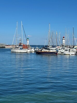 Un puerto deportivo con varios veleros amarrados, un faro al fondo y agua tranquila.