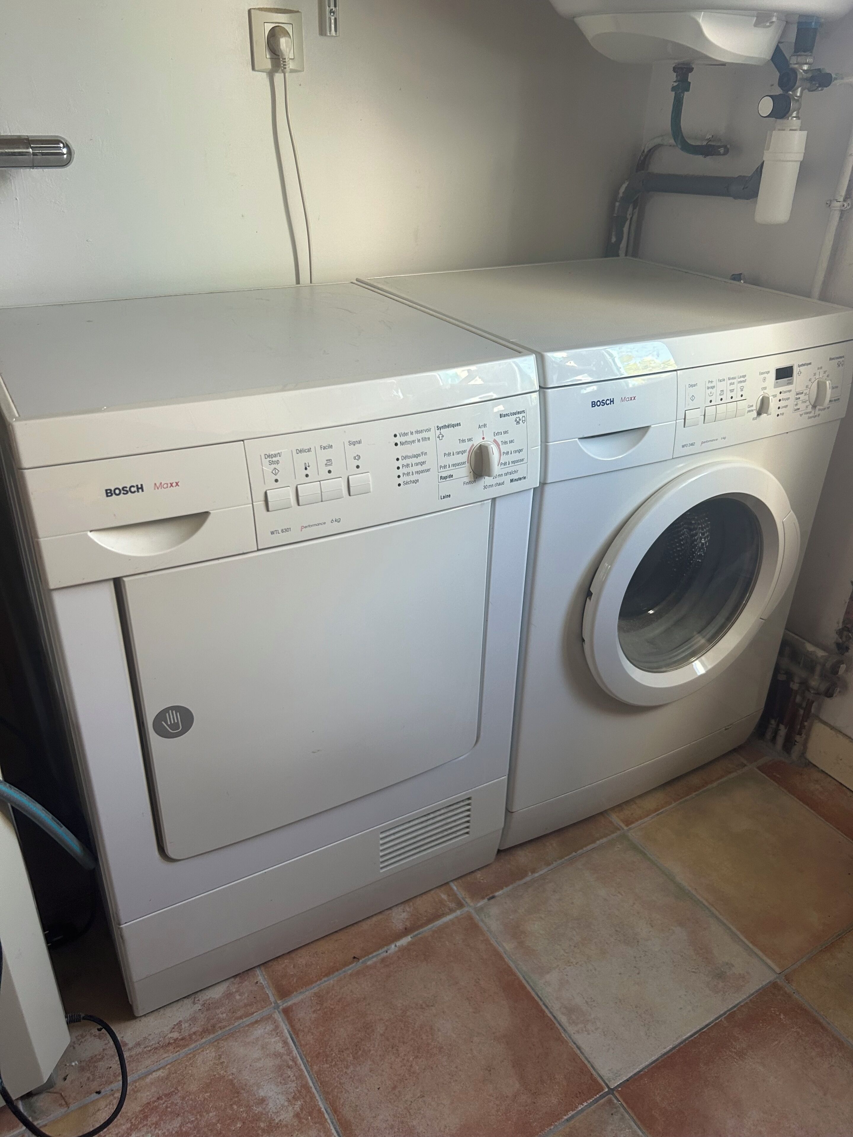 A white Bosch Maxx washing machine in a laundry room with tiled flooring and visible plumbing.