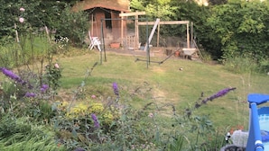 A garden with a variety of plants and flowers, a blue structure with pipes, and a wooden shed in the background.
