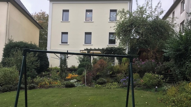 A grassy area with a swing set, surrounded by a garden with various plants and a building in the background.