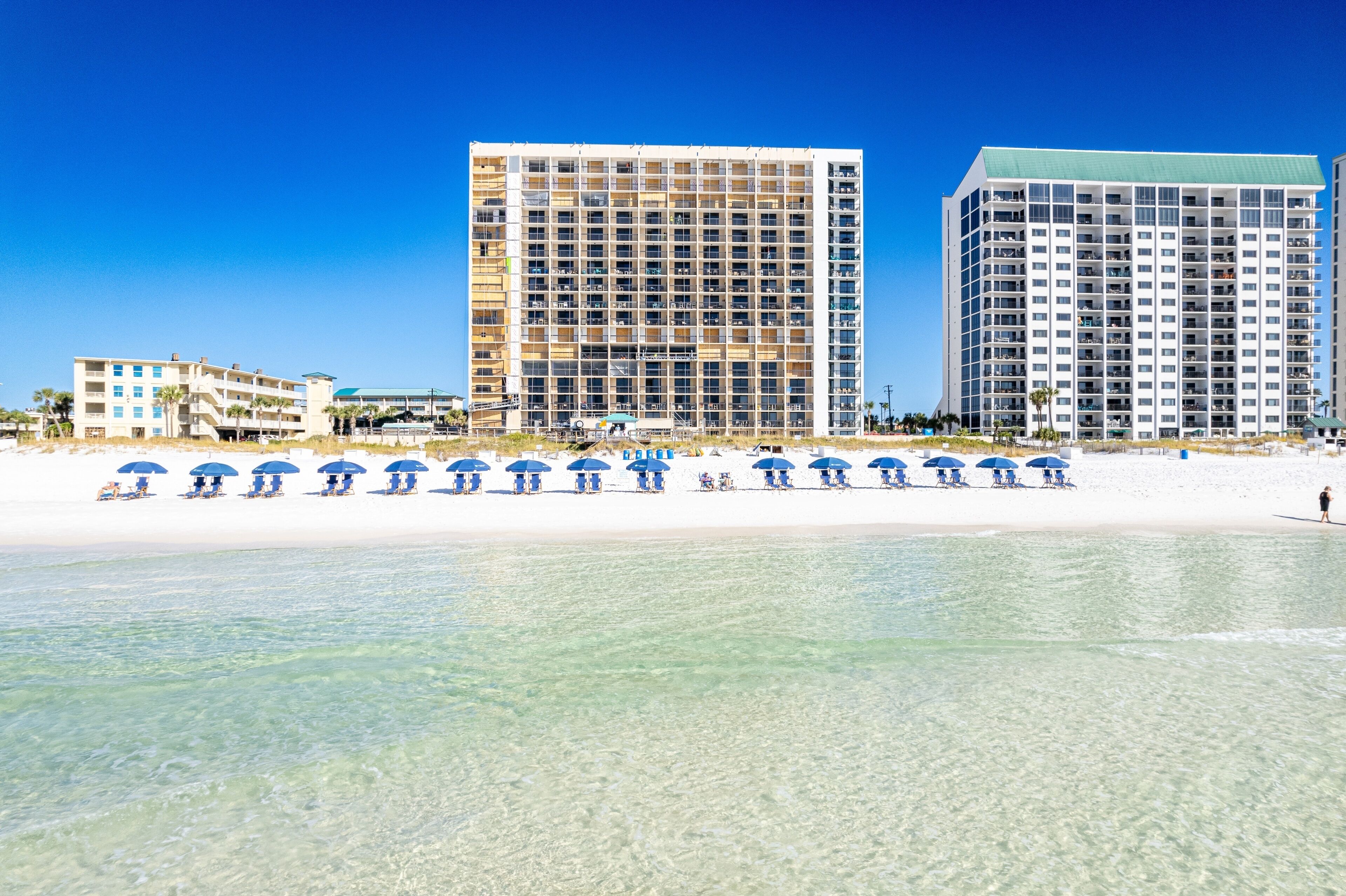 Un hotel frente a la playa con varios pisos, una playa de arena y varias sombrillas azules.