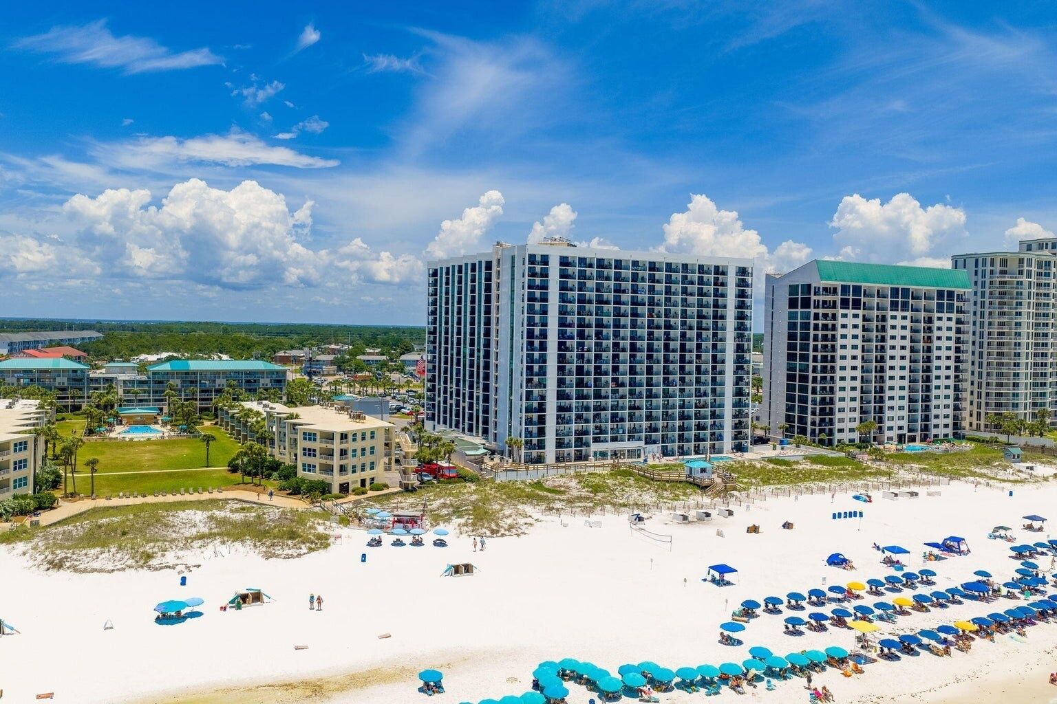 A beachfront hotel with multiple high-rise buildings, a sandy beach with umbrellas, and a clear blue sky.