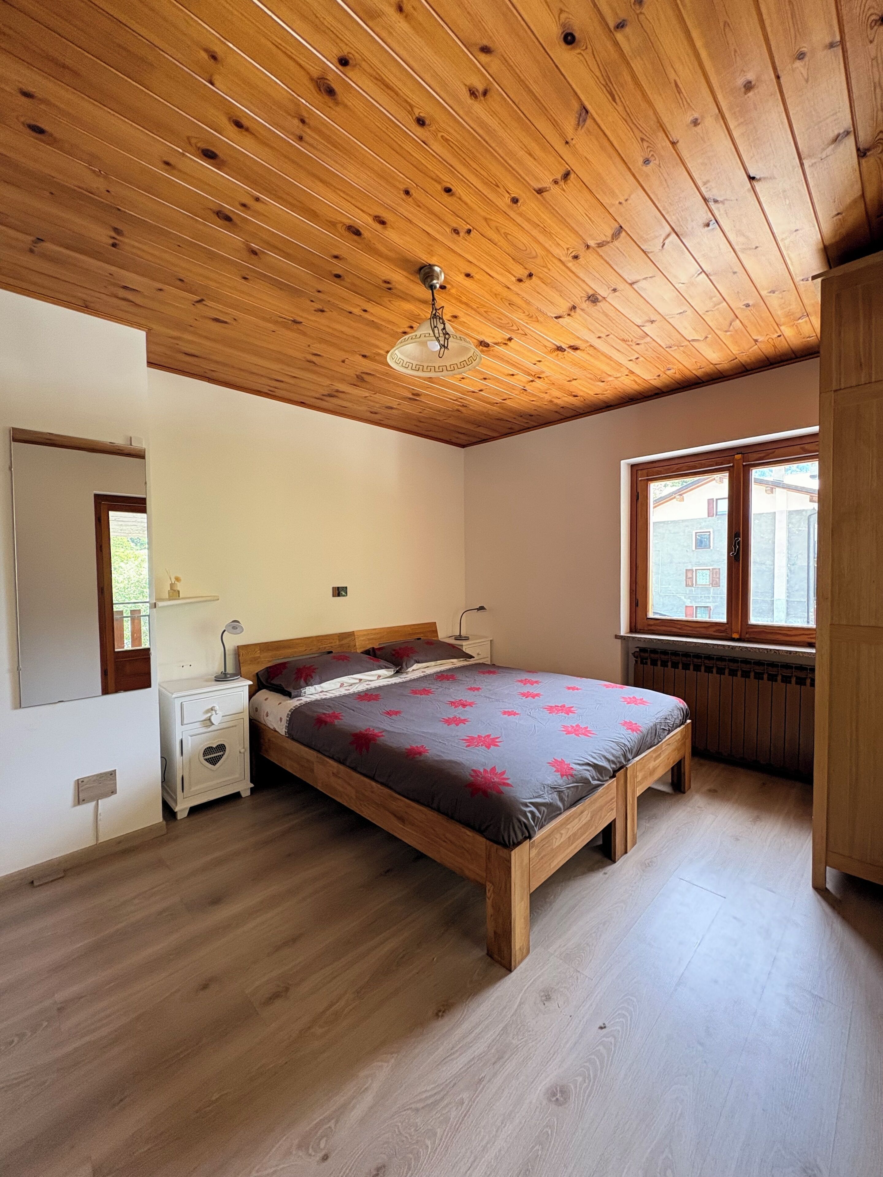 Un dormitorio con techo de madera, una cama con mesita de noche con forma de corazón, una ventana con vistas y un radiador.