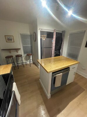 Eine kleine Küche mit Holzarbeitsplatte, einem eingebauten Backofen und einem Geschirrspüler. Es gibt Barhocker und ein Fenster mit Jalousien.