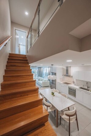 Une cuisine moderne avec des meubles blancs, un coin repas doté d’une table et de chaises blanches, et un escalier en bois menant à l’étage supérieur.