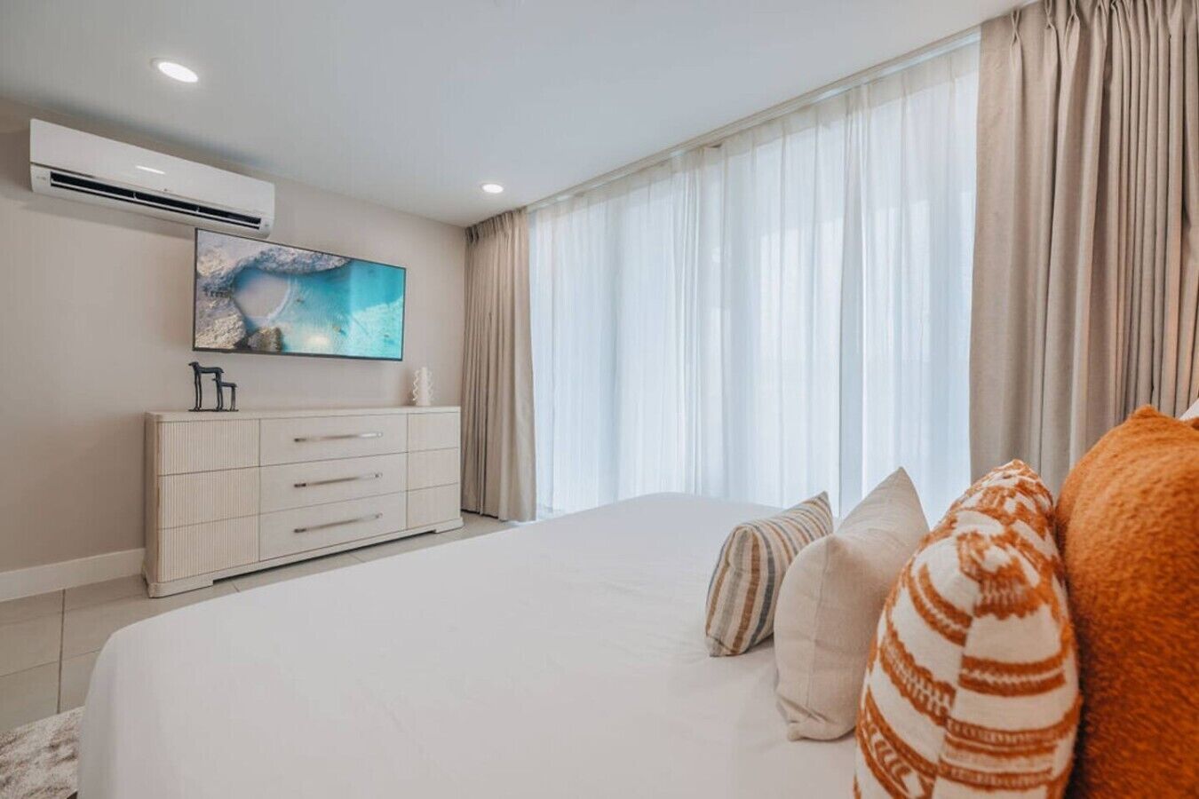 Un dormitorio con una cama grande, un aparador, un televisor de pantalla plana y un ventanal con cortinas.