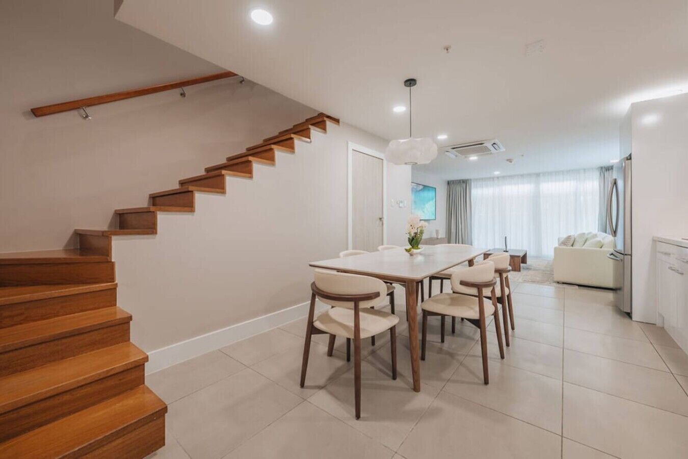 Un interior moderno con una escalera de madera, un comedor con mesa y sillas blancas, y una sala de estar con sofá y televisor.