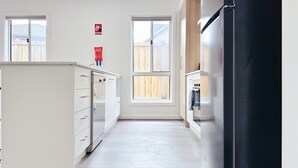 A kitchen with a refrigerator, stove, and cabinets, a fire blanket sign, and a window with a view of a wooden fence. - 2024Built/AC & Bath each Room/Study/TownCentre/SDA (Werribee)
