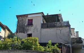 Ein Gebäude mit Balkon, einem Fenster mit roten Rollläden und blauem Himmel mit Vögeln im Hintergrund.