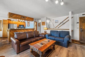 A living room with a brown leather sofa, a blue sofa, a wooden coffee table, a kitchen area with a refrigerator and microwave, and a wall with skis. - Phoenix 219 – Your Gondola-Side Mountain Retreat in Steamboat Springs! (Steamboat Springs)