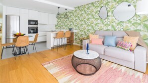 A modern kitchen with white cabinets, a dining area with wicker chairs, a round coffee table, and a living area with a sofa and patterned wallpaper. - Apartment in Las Palmas Near Playa Alcaravaneras (Las Palmas de Gran Canaria)