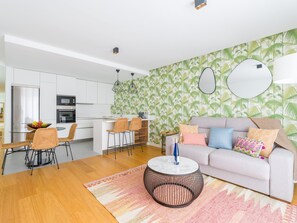 A modern kitchen with white cabinets, a dining area with wicker chairs, a round coffee table, and a living area with a sofa and patterned wallpaper. - Apartment in Las Palmas Near Playa Alcaravaneras (Las Palmas de Gran Canaria)