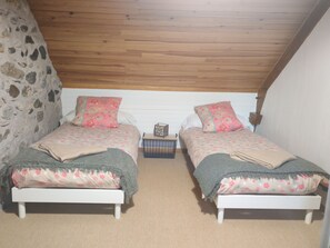 Quarto com duas camas, parede de pedra, vigas de madeira no teto e uma pequena mesa de cabeceira.