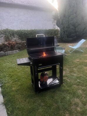 A black barbecue grill with a side table, a pair of grilling tools, and a bag of charcoal briquettes on the grass. - Mountain home, perfect for families (Aste-Béon)