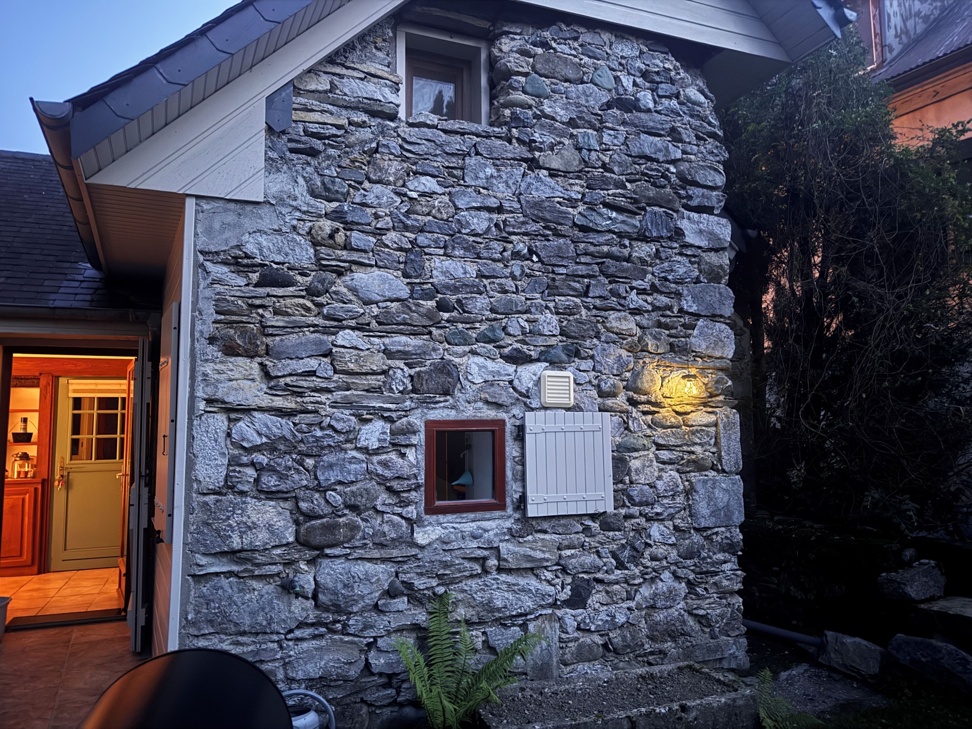A stone building with a sloped roof, a window with shutters, and a small vent on the wall.