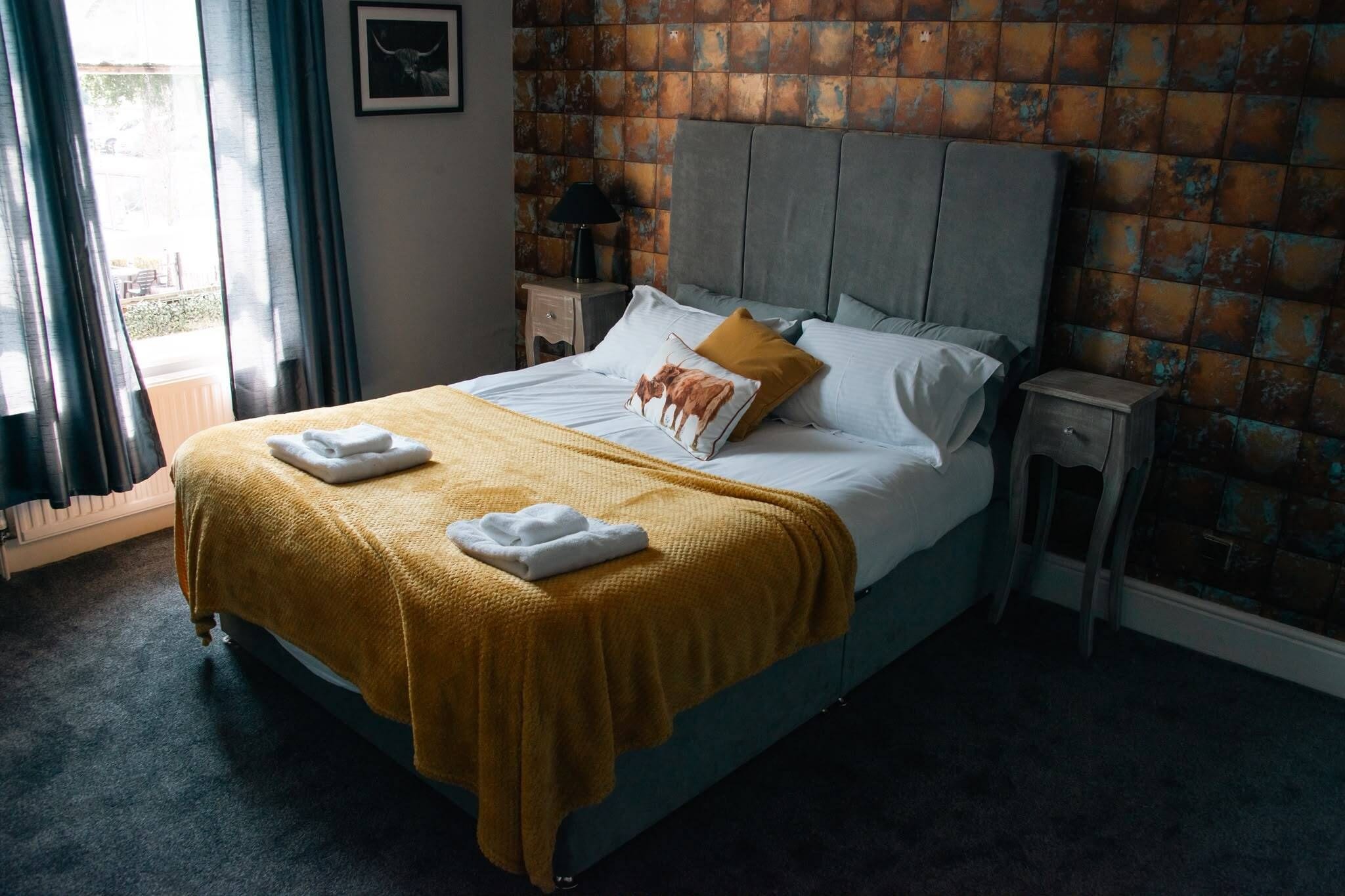 Una cama con cabecera, una mesita de noche, una lámpara y una ventana con cortinas.