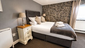 A neatly made bed with a dark headboard, a bedside table with a lamp, and a patterned wallpaper. - The Lobster Pots - Bawtry (Bawtry)