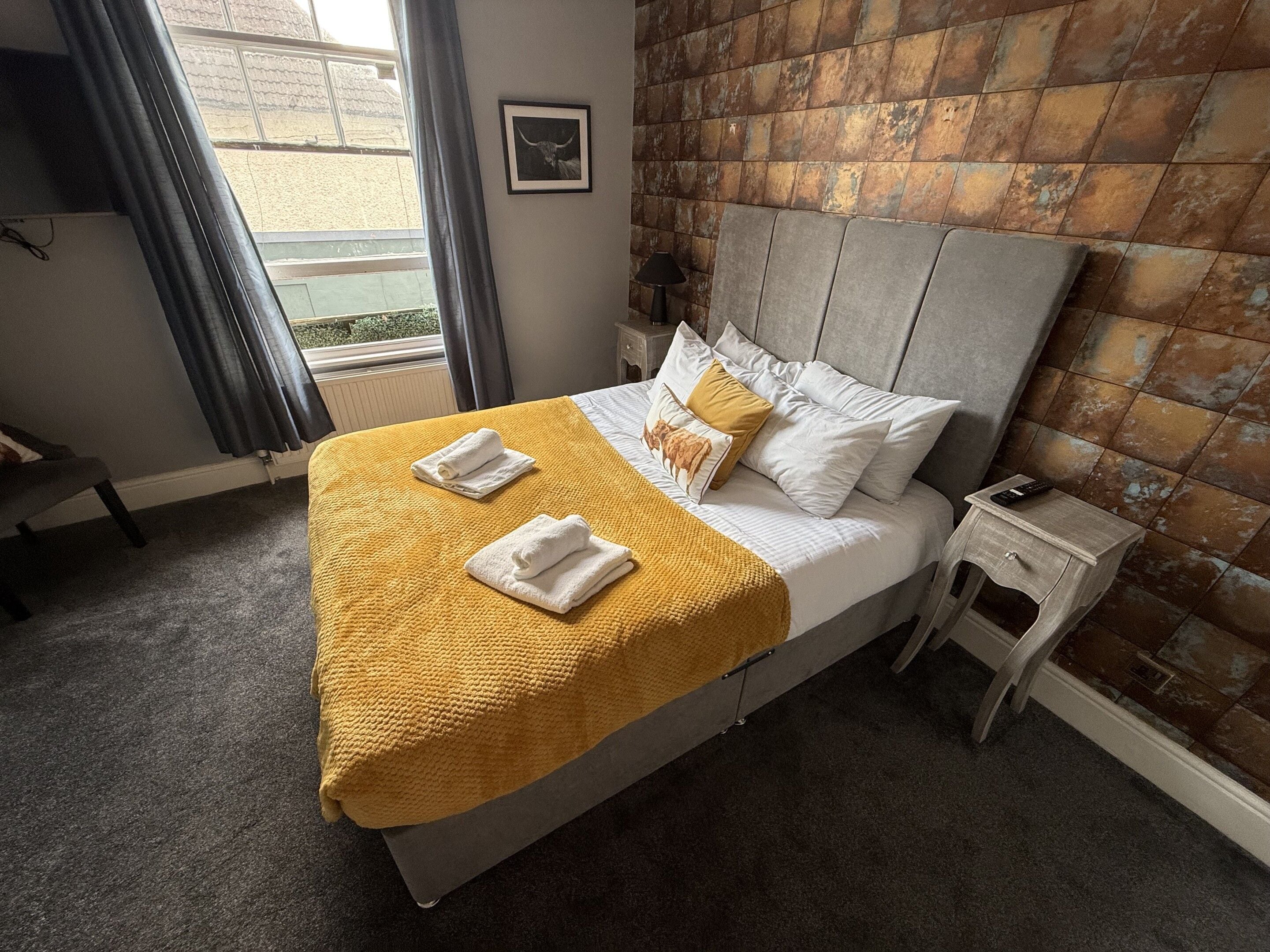 Un lit bien fait, avec une couverture jaune et deux serviettes, une table de chevet avec une télécommande et un mur décoré d’un motif en briques.
