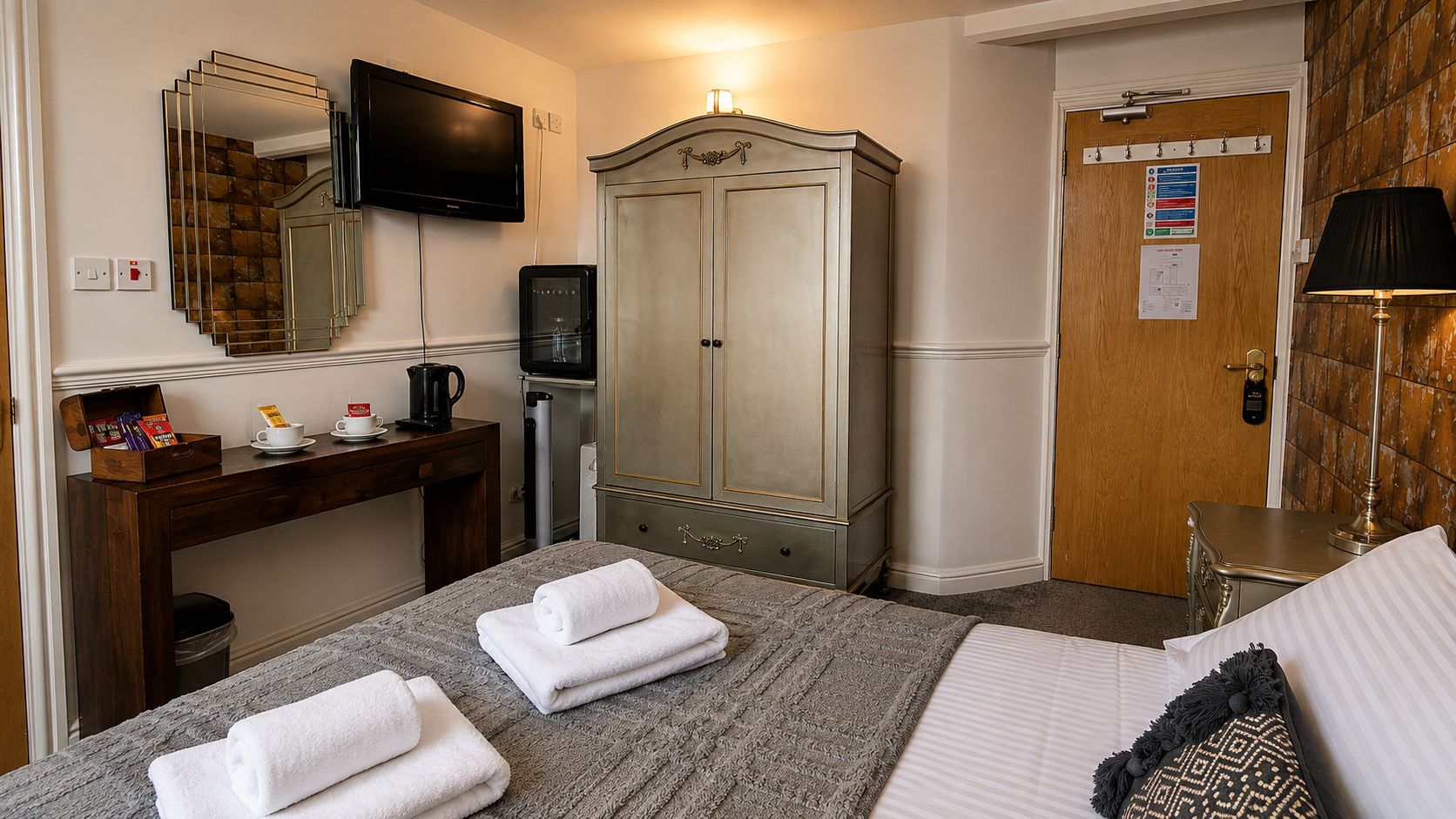 A hotel room with a bed, a desk with a coffee maker, a wardrobe, and a television mounted on the wall.