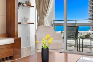 A modern living room with a sofa, a table with a vase of yellow flowers, a balcony with a view of the ocean, and a cityscape in the background. - Private Residence at Carillon Resort - 910 (Miami Beach)