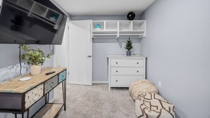 Quarto compacto com televisão, uma pequena planta, um penteou branco e um banco.