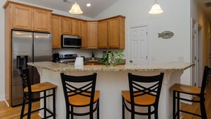 A kitchen with wooden cabinets, a stainless steel refrigerator, a microwave, and a granite countertop. There are three black chairs with wooden seats. - Inviting Single Family Home Near Beach - K403 (Kure Beach)