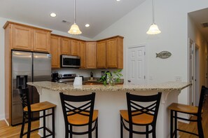 A kitchen with wooden cabinets, a stainless steel refrigerator, a microwave, and a granite countertop. There are three black chairs with wooden seats. - Inviting Single Family Home Near Beach - K403 (Kure Beach)