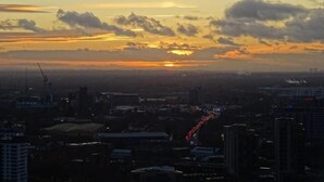 Paisagem urbana ao pôr do sol, com edifícios visíveis e uma estrada com veículos em movimento.