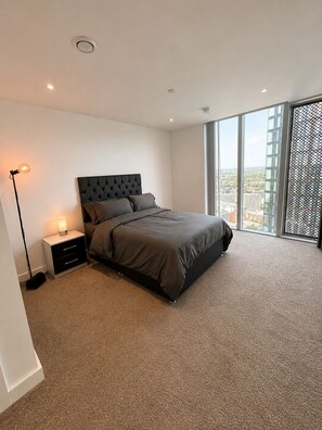 Quarto moderno com uma cama grande, uma mesinha de cabeceira, um abajur e vista para a cidade através de grandes janelas.