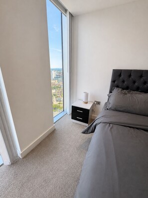 Ein Schlafzimmer mit großem Fenster, einem Bett, einem Nachttisch und Blick auf eine Stadtlandschaft.