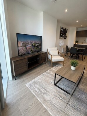 Ein modernes Wohnzimmer mit Flachbildfernseher, einer Couch, einem Essbereich mit Stühlen und einer Küche im Hintergrund.