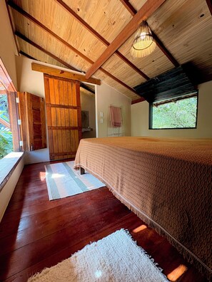 Ein Schlafzimmer mit Holzbalkendecke, einem Bett mit strukturierter Decke, einem Fenster mit Blick auf Bäume und einer Holztür.
