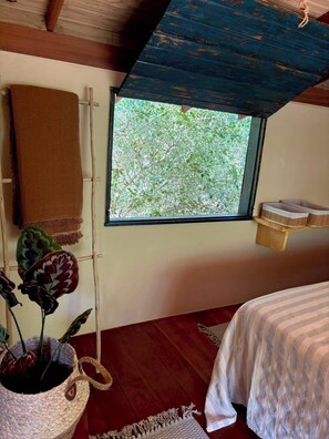 Ein Zimmer mit Holzboden, einem Fenster mit Blick auf Bäume, einem Bett mit gestreifter Bettdecke und einer Pflanze in einem gewobenen Korb.