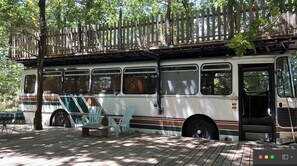A bus parked on a wooden deck with a wooden fence overhead, surrounded by trees. - Rooftop bus, outdoor shower + bathtub (dry toilet) (Mimizan)