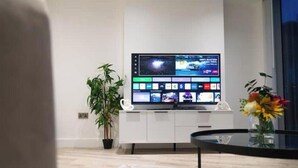 A modern living room with a flat-screen TV mounted on a white cabinet, a potted plant, a glass coffee table, and a vase with flowers. - Modern 2-Bed Penthouse in Deansgate | Stunning City Views (Manchester)