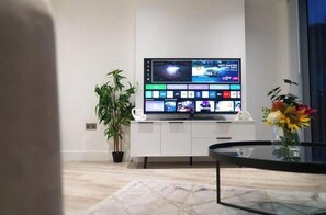 Ein modernes Wohnzimmer mit einem Flachbildfernseher an einem weißen Schrank, einer Topfpflanze, einem Glastisch und einer Vase mit Blumen.